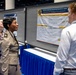 Three individuals are gathered at an academic or military research poster session, examining a research poster titled 'Stimulant Use, Screen Time, Sleep, and Fatigue in Deployed U.S. Sailors.' One person appears to be in a military uniform (tan/khaki), while the others are in civilian attire. The setting appears to be a large convention or conference hall with multiple poster displays.