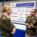 Two U.S. Army officers in dress green uniforms are engaged in conversation at what appears to be a military medical research conference or symposium. The officer on the right, identified by her name tag as 'Williams,' is presenting a research poster titled 'Automated Clinical Decision Support for Triage and Monitoring of Military Casualties: A Modified Delphi Consensus Study' sponsored by the Defense Health Agency (DHA). The poster displays research sections including Introduction, Methods, Results, and Conclusion with data visualizations.