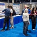 A scientific poster session at a professional conference, likely a medical or defense health research symposium. Attendees are gathered in small groups discussing research posters mounted on display boards. The venue features blue carpeting and blue-skirted display tables in a large convention hall.