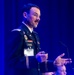 A U.S. Army officer in dress uniform is delivering a presentation or speech at what appears to be a formal conference or symposium. The officer, identified by his name tag and uniform name tape as Levi Jackson, is gesturing with his hands and holding what appears to be a presentation clicker. The setting features blue stage lighting against a dark curtain backdrop.