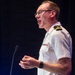 A military officer in a white uniform is speaking at a podium or lectern, appearing to be delivering a presentation or speech. He is wearing glasses and has rank insignia on his shoulder epaulette along with service ribbons on his chest. The image is taken from a side profile angle against a dark background.