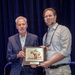 Two men are posing together at what appears to be a professional conference or symposium, jointly holding a wooden award plaque. The plaque reads 'BEST IN SHOW POSTER' and features an engraved military-themed image. The man on the right is wearing a conference name badge identifying him as 'W. BRETT JOHNSON'.
