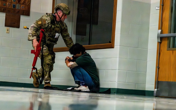A person in military/tactical gear and helmet, carrying a red training weapon, is leaning toward a civilian who is crouched against a wall in what appears to be a school or institutional hallway. The red-colored firearm indicates this is a training exercise or drill, not a live operation. The highly polished floor creates a vivid mirror reflection of both individuals.