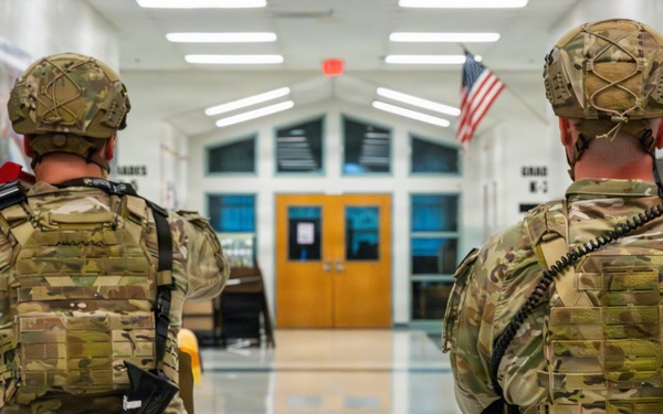 Two individuals in full military-style camouflage uniforms and tactical gear are standing in what appears to be a school hallway or lobby. They are carrying rifles fitted with blue training/simulation magazines, indicating this is a training exercise rather than a live operation. A U.S. flag is visible in the background near the entrance doors, and a banner reading 'WE ARE SCOTT' is partially visible on the left wall.