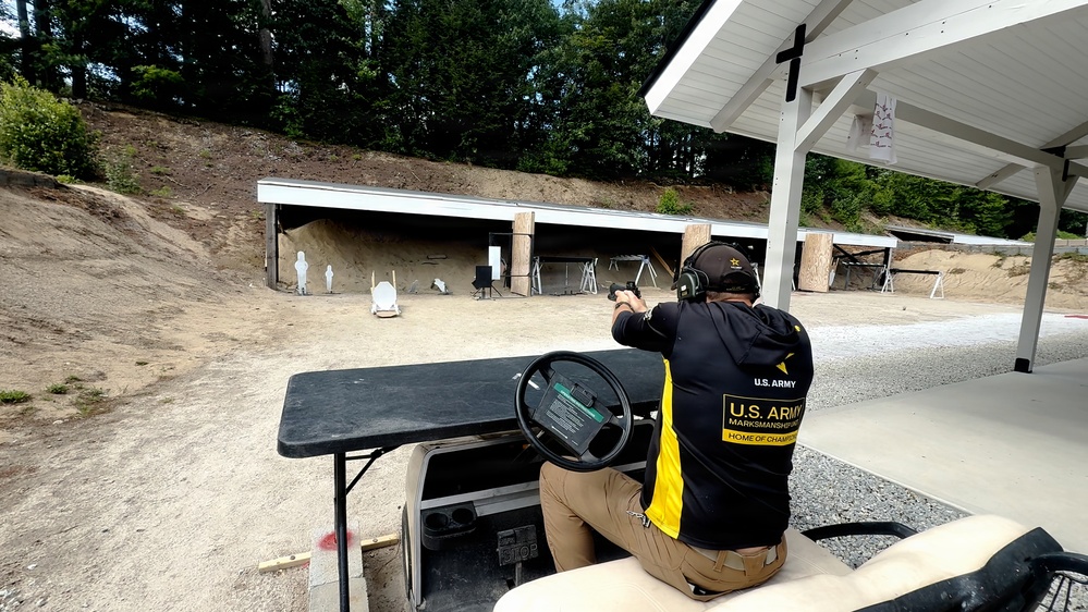 A U.S. Army Marksmanship Unit member is firing a pistol at an outdoor shooting range. The shooter is positioned at a firing line near a covered pavilion area, aiming downrange at multiple white silhouette targets set against a sandy berm. The facility appears to be a professional military or competitive shooting range.