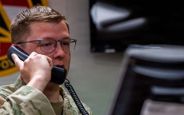 A U.S. Air Force enlisted member, identifiable by the 'U.S. AIR FORCE' tape and Technical Sergeant rank insignia on his OCP uniform, is seated at a workstation speaking on a corded telephone. He is wearing glasses and appears focused on a computer monitor in front of him. The setting appears to be a military operations or command center environment.
