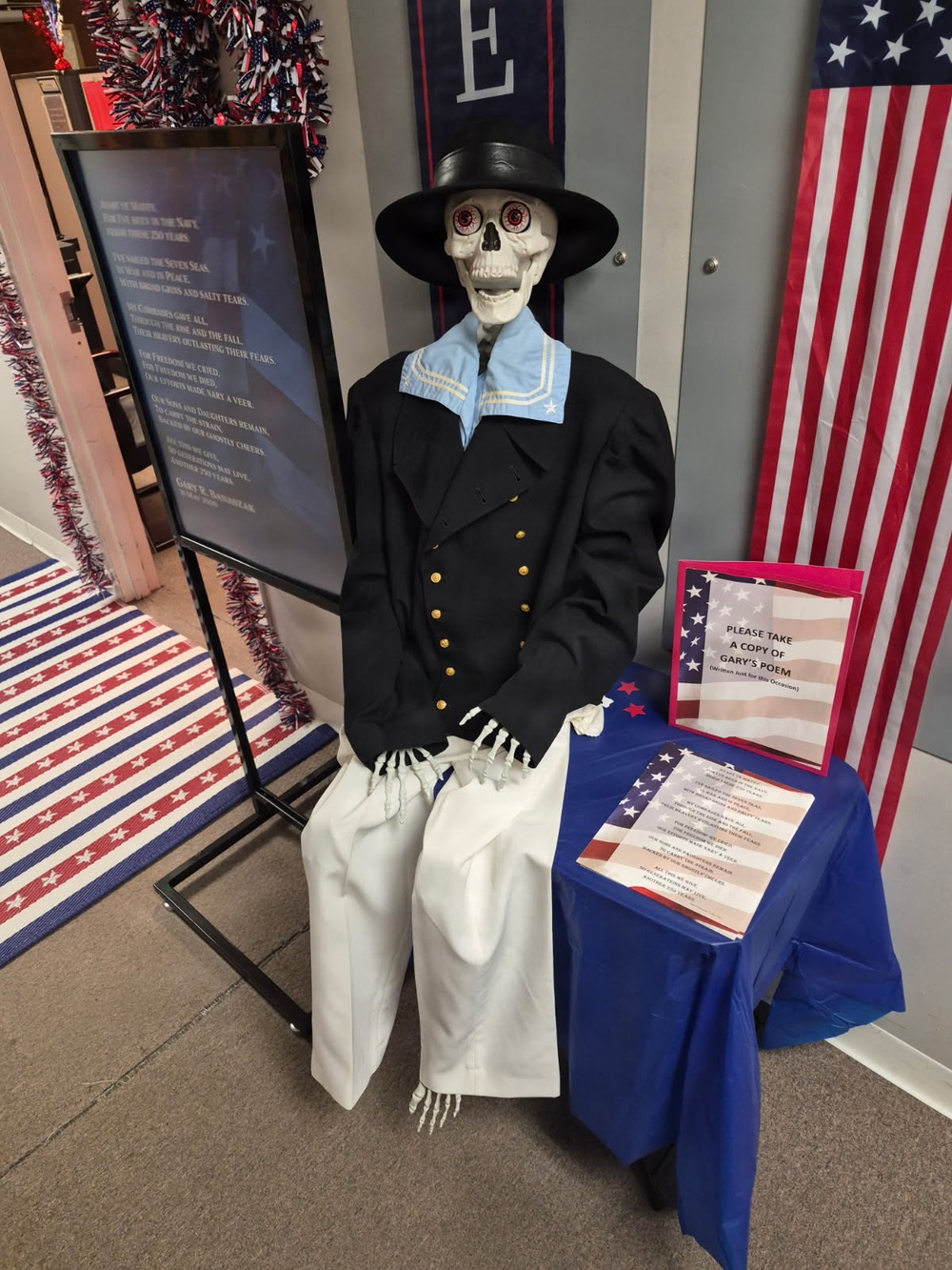 A decorative skeleton figure dressed in a Navy-style uniform with a black jacket, white trousers, sailor collar, and black top hat is displayed in what appears to be a patriotic-themed indoor setting. The display includes a framed poem on a stand to the left, attributed to 'Gary R. Bauslaur,' and a blue-draped table to the right with printed copies of the poem available for visitors to take. The overall setting features American patriotic decorations including U.S. flags, red-white-and-blue garland, and a striped rug.