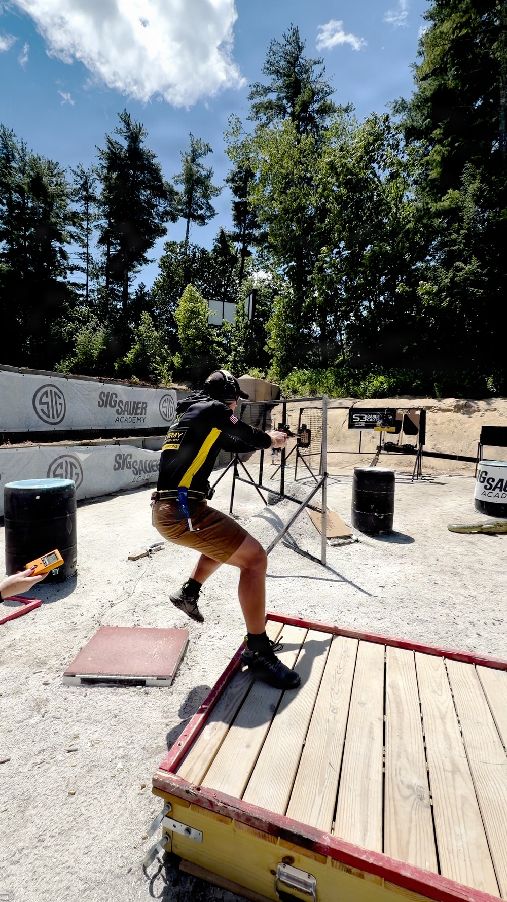 A competitive shooter is actively engaging targets at the SIG Sauer Academy shooting range. The shooter is in a dynamic stance on a wooden platform, aiming a firearm downrange toward steel targets. A timer operator is visible at the left edge of the frame, indicating this is a timed competitive shooting stage.