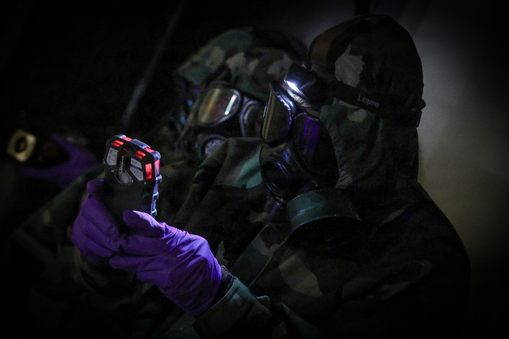 Two individuals wearing full CBRN (Chemical, Biological, Radiological, Nuclear) protective gear, including gas masks and camouflage suits, are shown in a low-light environment. The foreground figure is holding a small electronic detection device with red indicator lights while wearing purple nitrile gloves. The scene appears to depict a military or emergency response training or operational scenario involving hazardous material detection.