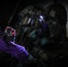 Two individuals wearing full CBRN (Chemical, Biological, Radiological, Nuclear) protective gear, including gas masks and camouflage suits, are shown in a low-light environment. The foreground figure is holding a small electronic detection device with red indicator lights while wearing purple nitrile gloves. The scene appears to depict a military or emergency response training or operational scenario involving hazardous material detection.