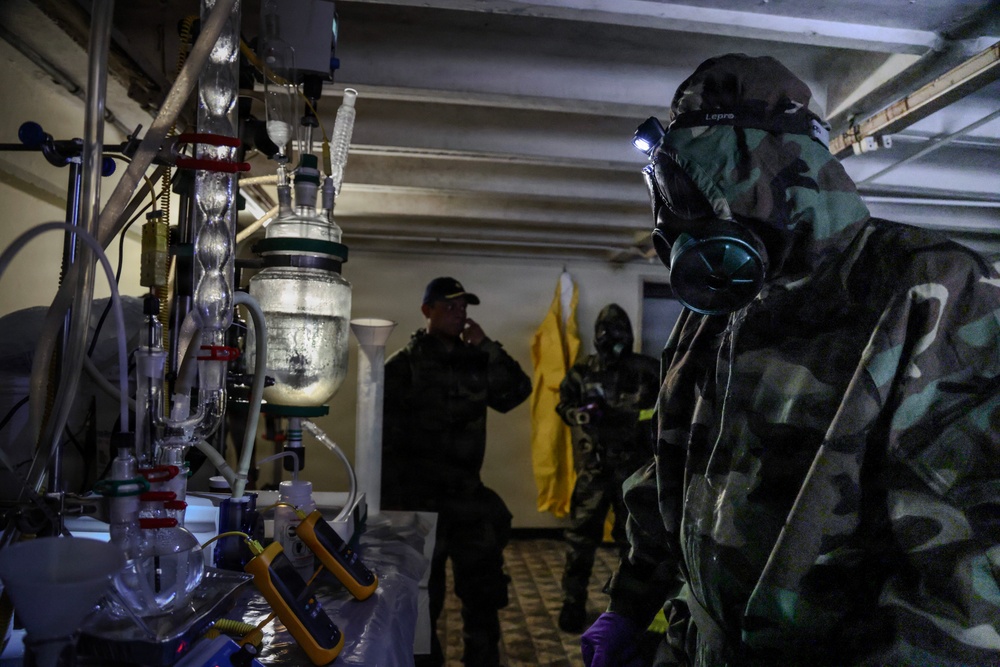 The image shows personnel in protective gear examining what appears to be a clandestine chemical laboratory. At least three individuals are present, with one wearing full CBRN (Chemical, Biological, Radiological, Nuclear) protective equipment including a gas mask and camouflage hazmat suit. The setting appears to be a law enforcement or military raid/inspection of an illicit drug or chemical production facility.
