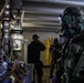 The image shows personnel in protective gear examining what appears to be a clandestine chemical laboratory. At least three individuals are present, with one wearing full CBRN (Chemical, Biological, Radiological, Nuclear) protective equipment including a gas mask and camouflage hazmat suit. The setting appears to be a law enforcement or military raid/inspection of an illicit drug or chemical production facility.