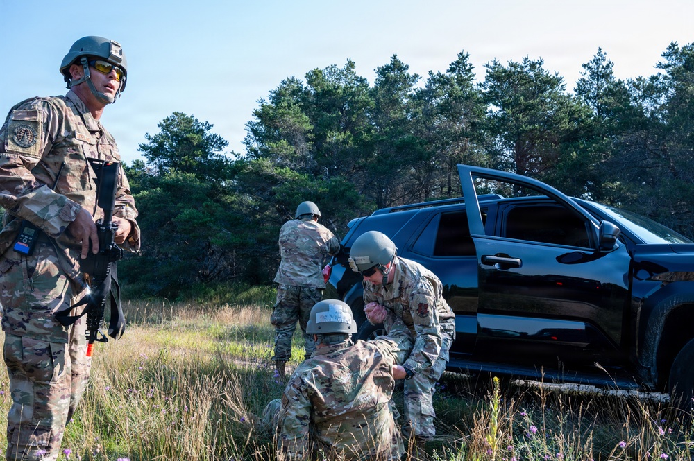 Four military personnel in MultiCam/OCP camouflage uniforms and ballistic helmets are conducting what appears to be a field training exercise near a black SUV with its doors open. One soldier stands guard with a rifle while the others appear to be performing a tactical casualty care or vehicle search scenario in a wooded, grassy outdoor environment. The scene is consistent with a military training exercise rather than a combat situation.
