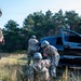 Four military personnel in MultiCam/OCP camouflage uniforms and ballistic helmets are conducting what appears to be a field training exercise near a black SUV with its doors open. One soldier stands guard with a rifle while the others appear to be performing a tactical casualty care or vehicle search scenario in a wooded, grassy outdoor environment. The scene is consistent with a military training exercise rather than a combat situation.