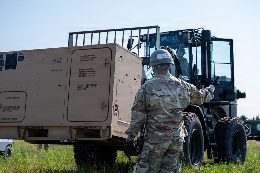 A military service member in OCP (Operational Camouflage Pattern) uniform and helmet is directing a Caterpillar forklift operator during a field operation. The forklift appears to be maneuvering a large tan military generator or equipment unit in an outdoor grassy area. This appears to be a training or logistics exercise involving heavy equipment operations.