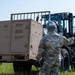 A military service member in OCP (Operational Camouflage Pattern) uniform and helmet is directing a Caterpillar forklift operator during a field operation. The forklift appears to be maneuvering a large tan military generator or equipment unit in an outdoor grassy area. This appears to be a training or logistics exercise involving heavy equipment operations.