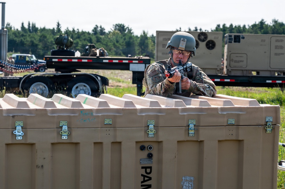 A soldier in full combat gear, including a helmet and protective eyewear, is taking an aiming position behind large tan military storage containers during what appears to be a training exercise. The weapon appears to have a red training device attached to the muzzle, indicating a simulated force-on-force exercise. The setting is an outdoor military installation with trailers, vehicles, and tree line visible in the background.