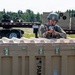 A soldier in full combat gear, including a helmet and protective eyewear, is taking an aiming position behind large tan military storage containers during what appears to be a training exercise. The weapon appears to have a red training device attached to the muzzle, indicating a simulated force-on-force exercise. The setting is an outdoor military installation with trailers, vehicles, and tree line visible in the background.