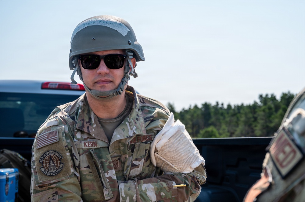 A U.S. Air Force service member named Kern is photographed in an outdoor field environment. He is wearing OCP (Operational Camouflage Pattern) uniform, a tactical helmet, and sunglasses, with his name tape and unit patch clearly visible. The setting appears to be a training or operational exercise near a vehicle.