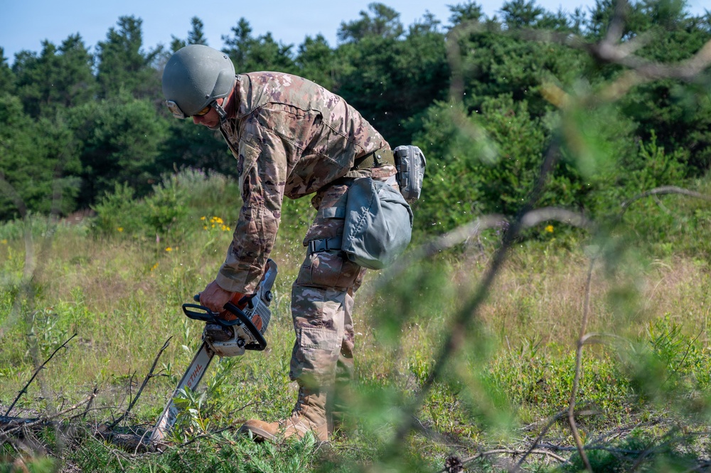 A military service member in full camouflage uniform and combat helmet is operating a STIHL chainsaw in an open field or clearing. The individual appears to be conducting land clearing or obstacle removal operations in a wooded outdoor environment. The scene suggests a military engineering or field operations task.