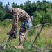 A military service member in full camouflage uniform and combat helmet is operating a STIHL chainsaw in an open field or clearing. The individual appears to be conducting land clearing or obstacle removal operations in a wooded outdoor environment. The scene suggests a military engineering or field operations task.