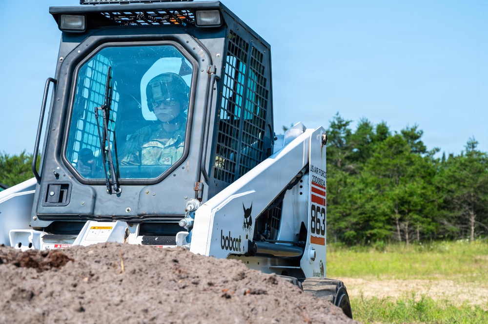 A military operator is driving a Bobcat 863 skid-steer loader marked with 'US Air Force' insignia and an identification number. The machine is actively moving a pile of dirt or soil in an outdoor field setting. The operator is wearing military-style protective gear including a helmet.