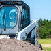 A military operator is driving a Bobcat 863 skid-steer loader marked with 'US Air Force' insignia and an identification number. The machine is actively moving a pile of dirt or soil in an outdoor field setting. The operator is wearing military-style protective gear including a helmet.