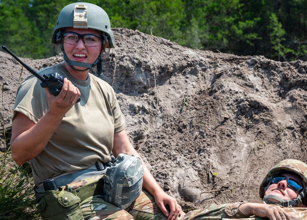 Two military personnel are positioned near a large earthen berm or dirt mound in an outdoor training environment. A female service member in the foreground is wearing a combat helmet and protective eyewear while holding a handheld radio, appearing to communicate. A second individual in camouflage uniform and sunglasses is lying on the ground beside her.