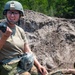 Two military personnel are positioned near a large earthen berm or dirt mound in an outdoor training environment. A female service member in the foreground is wearing a combat helmet and protective eyewear while holding a handheld radio, appearing to communicate. A second individual in camouflage uniform and sunglasses is lying on the ground beside her.