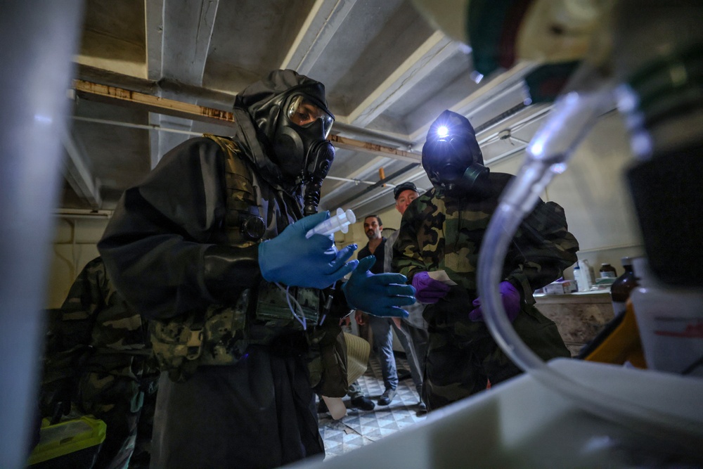 Two individuals wearing full CBRN (Chemical, Biological, Radiological, Nuclear) protective gear including gas masks, camouflage suits, and blue nitrile gloves are handling a syringe in what appears to be an indoor basement or low-ceiling room. A third person in civilian clothing stands in the background observing. The setting and equipment suggest a hazmat response, military training exercise, or decontamination operation.