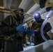 Two individuals wearing full CBRN (Chemical, Biological, Radiological, Nuclear) protective gear including gas masks, camouflage suits, and blue nitrile gloves are handling a syringe in what appears to be an indoor basement or low-ceiling room. A third person in civilian clothing stands in the background observing. The setting and equipment suggest a hazmat response, military training exercise, or decontamination operation.