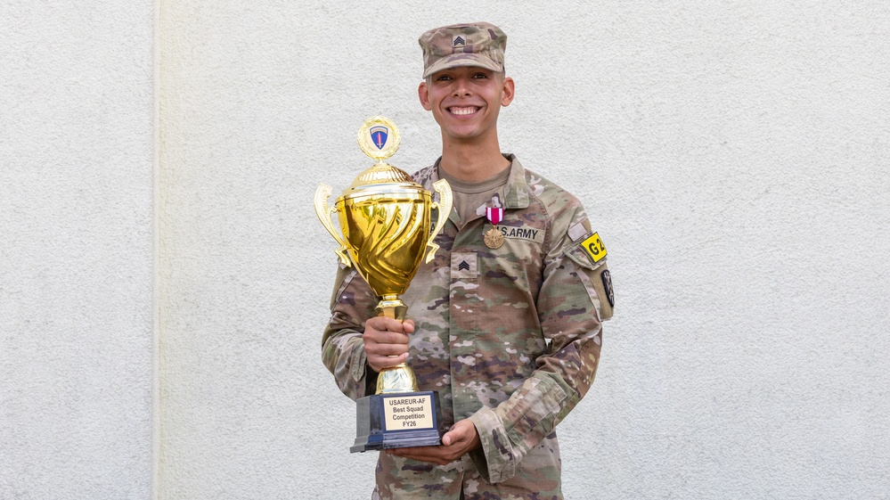 A U.S. Army Sergeant in Army Combat Uniform (ACU/OCP camouflage) poses proudly holding a large gold trophy. The trophy's base plate reads 'USAREUR-AF Best Squad Competition FY26'. The soldier is wearing a medal on their chest and smiling broadly against a light-colored wall background.
