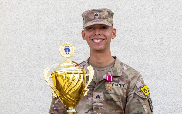 A U.S. Army Sergeant in Army Combat Uniform (ACU/OCP camouflage) poses proudly holding a large gold trophy. The trophy's base plate reads 'USAREUR-AF Best Squad Competition FY26'. The soldier is wearing a medal on their chest and smiling broadly against a light-colored wall background.