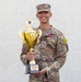 A U.S. Army Sergeant in Army Combat Uniform (ACU/OCP camouflage) poses proudly holding a large gold trophy. The trophy's base plate reads 'USAREUR-AF Best Squad Competition FY26'. The soldier is wearing a medal on their chest and smiling broadly against a light-colored wall background.