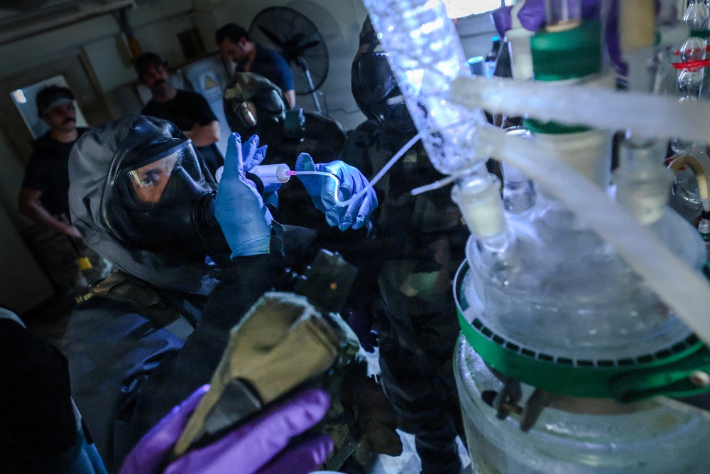 Personnel wearing full protective gear including gas masks, face shields, and chemical-resistant gloves are working around what appears to be a large chemical reactor or processing vessel. The scene suggests a hazardous materials operation, possibly a law enforcement drug lab raid, clandestine lab dismantlement, or HAZMAT training exercise. Multiple individuals in varying levels of protective equipment are visible in a confined indoor space.