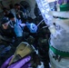 Personnel wearing full protective gear including gas masks, face shields, and chemical-resistant gloves are working around what appears to be a large chemical reactor or processing vessel. The scene suggests a hazardous materials operation, possibly a law enforcement drug lab raid, clandestine lab dismantlement, or HAZMAT training exercise. Multiple individuals in varying levels of protective equipment are visible in a confined indoor space.