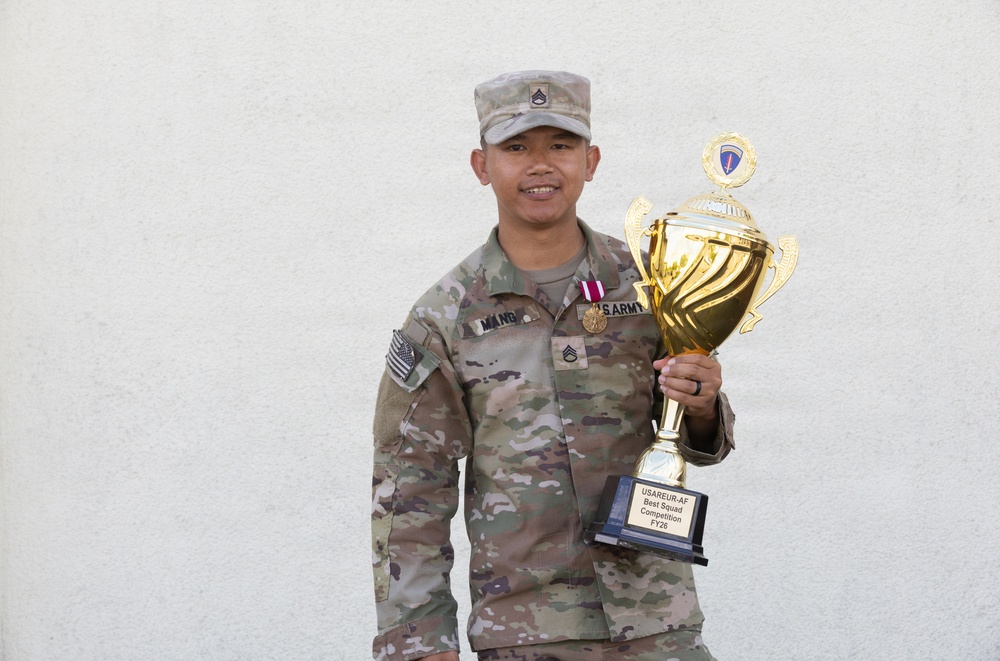 A U.S. Army soldier in MultiCam/OCP uniform is posing with a large gold trophy against a white wall. The soldier, identified by nametape as 'MANG,' holds the USAREUR-AF Best Squad Competition FY26 trophy. The soldier is smiling and appears to be celebrating a competitive achievement.
