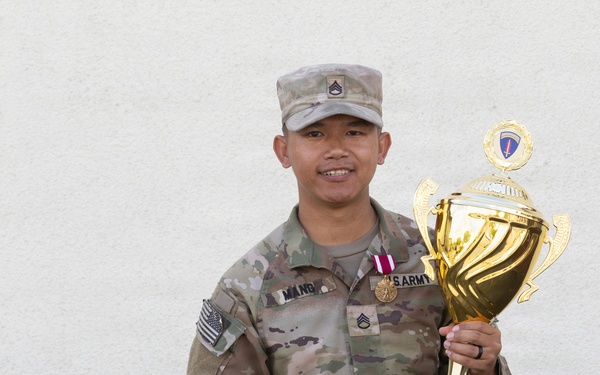 A U.S. Army soldier in MultiCam/OCP uniform is posing with a large gold trophy against a white wall. The soldier, identified by nametape as 'MANG,' holds the USAREUR-AF Best Squad Competition FY26 trophy. The soldier is smiling and appears to be celebrating a competitive achievement.
