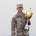 A U.S. Army soldier in MultiCam/OCP uniform is posing with a large gold trophy against a white wall. The soldier, identified by nametape as 'MANG,' holds the USAREUR-AF Best Squad Competition FY26 trophy. The soldier is smiling and appears to be celebrating a competitive achievement.