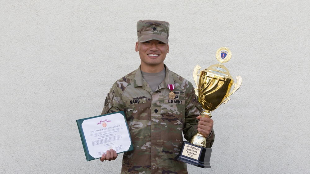 A U.S. Army soldier identified by name tape as 'MANG' poses proudly holding a large gold trophy and a certificate. He is wearing OCP (Operational Camouflage Pattern) uniform with a meritorious service medal ribbon and U.S. Army tape visible. The trophy inscription indicates he won the USAREUR-AF Best Squad Competition FY26.