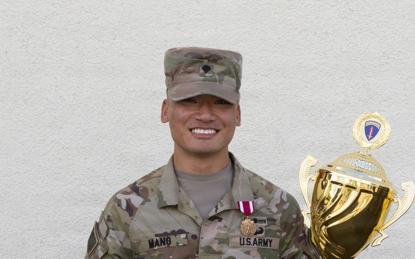 A U.S. Army soldier identified by name tape as 'MANG' poses proudly holding a large gold trophy and a certificate. He is wearing OCP (Operational Camouflage Pattern) uniform with a meritorious service medal ribbon and U.S. Army tape visible. The trophy inscription indicates he won the USAREUR-AF Best Squad Competition FY26.