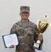 A U.S. Army soldier identified by name tape as 'MANG' poses proudly holding a large gold trophy and a certificate. He is wearing OCP (Operational Camouflage Pattern) uniform with a meritorious service medal ribbon and U.S. Army tape visible. The trophy inscription indicates he won the USAREUR-AF Best Squad Competition FY26.