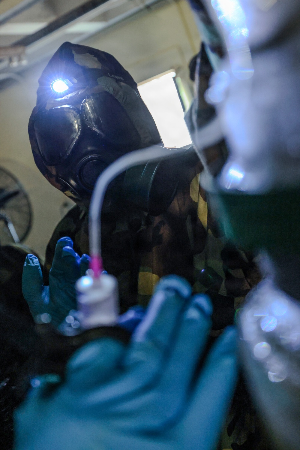 A person wearing a military-style gas mask with an attached headlamp and camouflage uniform is performing what appears to be a medical or technical procedure in a low-light environment. They are wearing blue nitrile gloves and handling medical tubing or IV line components. The image is taken from a close, low angle with bokeh effects in the foreground.