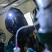 A person wearing a military-style gas mask with an attached headlamp and camouflage uniform is performing what appears to be a medical or technical procedure in a low-light environment. They are wearing blue nitrile gloves and handling medical tubing or IV line components. The image is taken from a close, low angle with bokeh effects in the foreground.