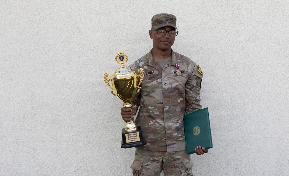 A U.S. Army soldier in OCP (Operational Camouflage Pattern) uniform poses against a white stucco wall holding a gold trophy and a green certificate folder. The trophy's base is inscribed with 'USAREUR-AF Best Squad Competition FY2X', indicating the soldier won a military competition. The soldier is wearing a medal on their chest and displays rank insignia and unit patches.