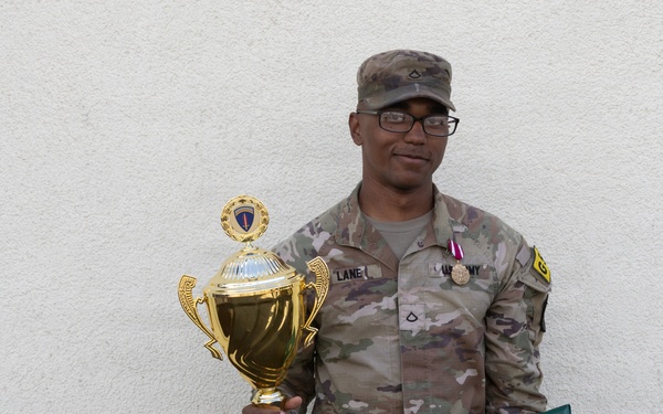 A U.S. Army soldier in OCP (Operational Camouflage Pattern) uniform poses against a white stucco wall holding a gold trophy and a green certificate folder. The trophy's base is inscribed with 'USAREUR-AF Best Squad Competition FY2X', indicating the soldier won a military competition. The soldier is wearing a medal on their chest and displays rank insignia and unit patches.