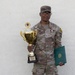 A U.S. Army soldier in OCP (Operational Camouflage Pattern) uniform poses against a white stucco wall holding a gold trophy and a green certificate folder. The trophy's base is inscribed with 'USAREUR-AF Best Squad Competition FY2X', indicating the soldier won a military competition. The soldier is wearing a medal on their chest and displays rank insignia and unit patches.