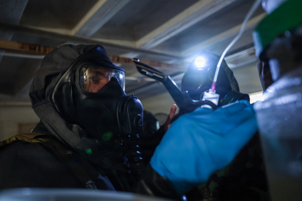 Two individuals wearing full-face gas masks and protective gear are working in a confined, dimly lit space, likely a crawl space or enclosed structure. One person is equipped with a mounted flashlight/headlamp that illuminates the area. The scene suggests a hazardous materials operation, emergency response drill, or inspection in a potentially contaminated environment.