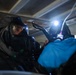 Two individuals wearing full-face gas masks and protective gear are working in a confined, dimly lit space, likely a crawl space or enclosed structure. One person is equipped with a mounted flashlight/headlamp that illuminates the area. The scene suggests a hazardous materials operation, emergency response drill, or inspection in a potentially contaminated environment.
