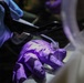 A close-up laboratory scene showing two gloved hands — one wearing a blue nitrile glove and one wearing a purple nitrile glove — performing a precise liquid transfer. A flexible tubing or pipette tip is being inserted into a small open glass vial held by the purple-gloved hand. The image is taken in low-light conditions with a shallow depth of field, suggesting a controlled laboratory or forensic environment.